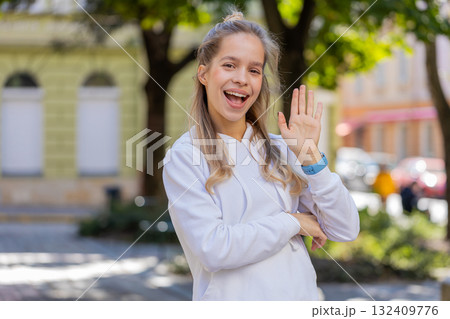 Cheerful young woman tourist smiling at camera, waving hands gesturing hello standing on city street Cheerful young woman tourist smiling at camera, waving hands gesturing hello standing on city street 132409776