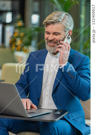 Mature businessman in suit answering to client on mobile phone call, using laptop in office lobby 132409917