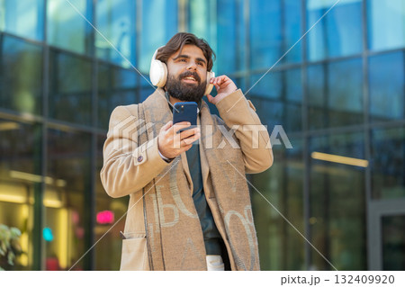 Happy young man putting on headphones listening favorite energetic music on smartphone on street 132409920