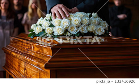 A person's hand resting on a wooden casket adorned with white roses during a funeral ceremony. A person's hand resting on a wooden casket adorned with white roses during a funeral ceremony. 132410920