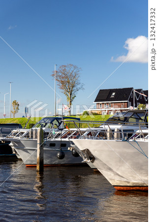 Modern luxury boats moored at Zwartsluis Netherlands canal marina bright sunlight modern houses trees background. Calm water reflects the vessels and blue sky, creating a serene Dutch coastal scene Modern luxury boats moored at Zwartsluis Netherlands canal marina bright sunlight modern houses trees background. Calm water reflects the vessels and blue sky, creating a serene Dutch coastal scene 132417332