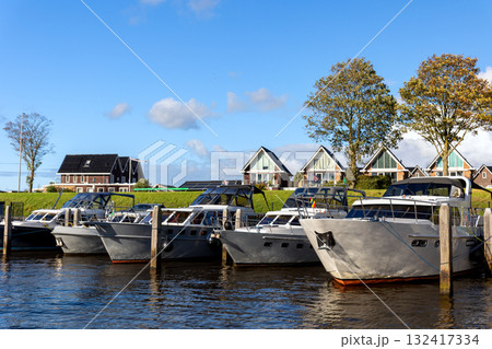 Modern luxury boats moored at Zwartsluis Netherlands canal marina bright sunlight modern houses trees background. Calm water reflects the vessels and blue sky, creating a serene Dutch coastal scene Modern luxury boats moored at Zwartsluis Netherlands canal marina bright sunlight modern houses trees background. Calm water reflects the vessels and blue sky, creating a serene Dutch coastal scene 132417334