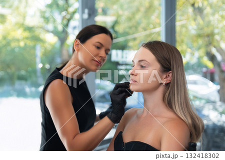 Makeup artist applies makeup to client in a bright studio with natural light and greenery outside Makeup artist applies makeup to client in a bright studio with natural light and greenery outside 132418302