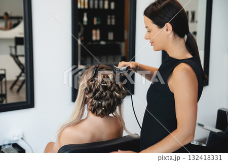 A hair stylist is curling a client hair with a styling tool in a contemporary salon. The setting is bright and organized, creating a stylish atmosphere for beauty services A hair stylist is curling a client hair with a styling tool in a contemporary salon. The setting is bright and organized, creating a stylish atmosphere for beauty services 132418331