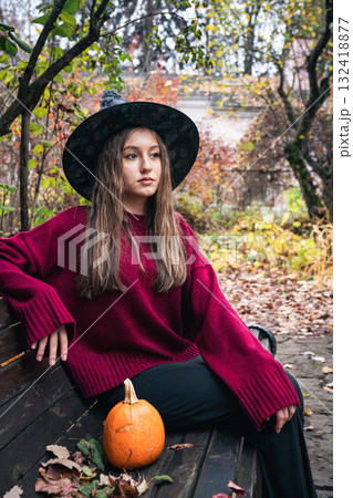 Young woman in a burgundy sweater and black hat sits on a park bench beside a small pumpkin, surrounded by autumn foliage 132418877