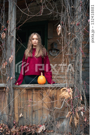 Young woman in a witch hat and red sweater stands near a rustic wooden house with a pumpkin on the railing, surrounded by dry vines 132418881