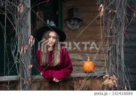 Young woman in a burgundy sweater stands on a rustic wooden porch with a pumpkin nearby, surrounded by autumn vines 132418889