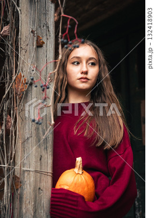 Young woman in a burgundy sweater holding a small pumpkin stands by a wooden porch covered with dry ivy 132418903