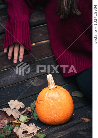 Close-up of a small orange pumpkin on a park bench next to a person in a burgundy sweater, surrounded by autumn leaves 132418904