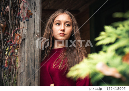 Young woman in a burgundy sweater stands by an old wooden porch surrounded by autumn foliage and ivy 132418905