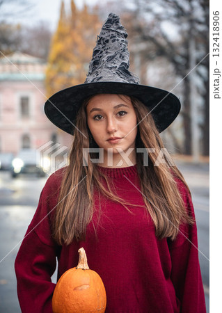 Young woman in a witch hat and burgundy sweater holding a pumpkin walks along an autumn city street. Halloween portrait 132418906