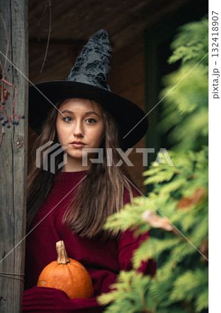 Young woman in a burgundy sweater stands on the wooden porch of an old house holding a small pumpkin, surrounded by autumn leaves and greenery Young woman in a burgundy sweater stands on the wooden porch of an old house holding a small pumpkin, surrounded by autumn leaves and greenery 132418907