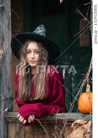Young woman in a witch hat and burgundy sweater leans out of a rustic wooden window surrounded by dry vines and a pumpkin, Halloween atmosphere Young woman in a witch hat and burgundy sweater leans out of a rustic wooden window surrounded by dry vines and a pumpkin, Halloween atmosphere 132418908