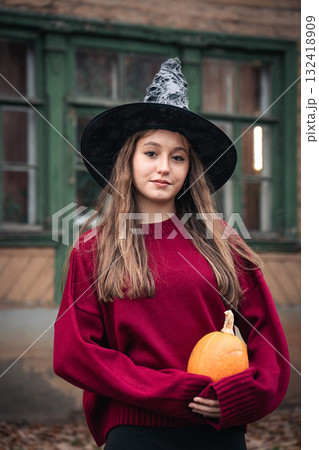 Young woman in a witch hat and burgundy sweater holds a pumpkin in front of an old wooden house with green window frames 132418909
