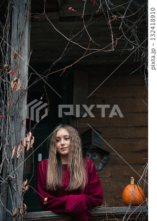 Young woman in a burgundy sweater stands on a rustic wooden porch with a pumpkin nearby, surrounded by autumn vines Young woman in a burgundy sweater stands on a rustic wooden porch with a pumpkin nearby, surrounded by autumn vines 132418910