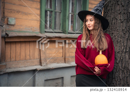 Young woman in a witch hat and burgundy sweater holds a pumpkin in front of an old wooden house with green window frames Young woman in a witch hat and burgundy sweater holds a pumpkin in front of an old wooden house with green window frames 132418914