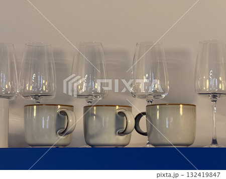 Ceramic mugs and wine glasses arranged on blue shelf. Kitchen interior, minimalist organization and aesthetic harmony of shapes, materials and reflections. 132419847