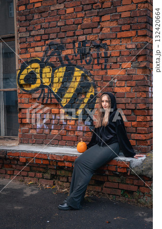 Teen girl in a black hoodie leans against a graffiti brick wall with a small pumpkin beside her Teen girl in a black hoodie leans against a graffiti brick wall with a small pumpkin beside her 132420664