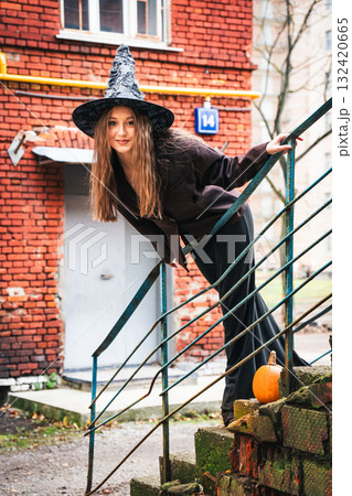 Smiling teen girl in a witch hat leans playfully on old stairs with a pumpkin nearby Smiling teen girl in a witch hat leans playfully on old stairs with a pumpkin nearby 132420665