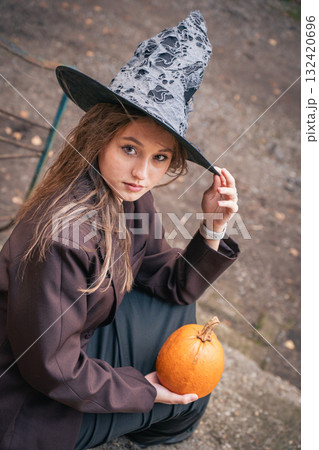 Smiling teen girl in a witch hat and brown blazer holds a small pumpkin outdoors 132420696