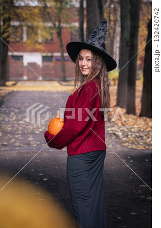 Teen girl in a witch hat and red sweater holding a pumpkin in a park alley with autumn leaves. Perfect for Halloween 132420742