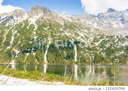 alpine lake in spring on a sunny day. travel landscape of rysy ridge in high tatra mountains of poland. snow among rocks and spruce forest on the hillside reflection in water. blue sky with clouds 132420769