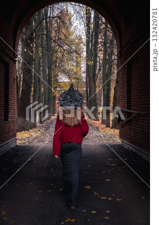 Teen girl in a witch hat and red sweater walking under an archway toward an autumn park with fallen leaves. Great for Halloween 132420781