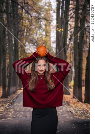 Teen girl in a red sweater holding a small pumpkin on her head in a park with autumn trees. Great for Halloween Teen girl in a red sweater holding a small pumpkin on her head in a park with autumn trees. Great for Halloween 132420787