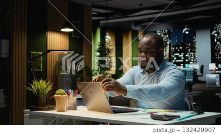 African american office worker placing a sticky note on his laptop as a reminder, trying to remember a task from his to do list. Working at night and leaving a memo for next morning. 132420907