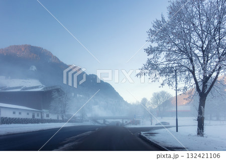 Scenic quiet winter road frosty valley covered mist fog and snow. Bare trees and distant mountains glow softly under the pale morning light. Alpine mountain village winter morning sunrise Scenic quiet winter road frosty valley covered mist fog and snow. Bare trees and distant mountains glow softly under the pale morning light. Alpine mountain village winter morning sunrise 132421106