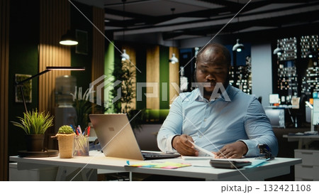 Livid black businessman breaking a paper sheet after making a mistake, handwriting and notetaking in office after hours. Displeased impatient employee redoing the draft on a new page. Livid black businessman breaking a paper sheet after making a mistake, handwriting and notetaking in office after hours. Displeased impatient employee redoing the draft on a new page. 132421108