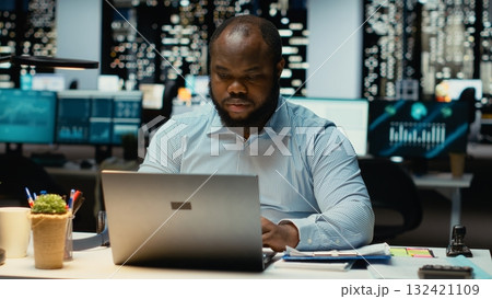 Black office worker reclining in a chair and relaxing at midnight, daydreaming during a work break. Fulfilled and self assured, reflecting on career success and corporate achievements. 132421109