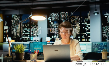 Businesswoman seeing the time as she concentrates on a complex task, finishing her idea and closing the lamp to leave to office after hours. Doing overtime in a dark office at midnight. Camera B. Businesswoman seeing the time as she concentrates on a complex task, finishing her idea and closing the lamp to leave to office after hours. Doing overtime in a dark office at midnight. Camera B. 132421158