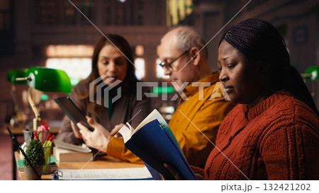 African american woman asking people for silence in a public library, being disturbed by their collective studying with a lot of noise. Mature colleagues learning with disrespect. 132421202