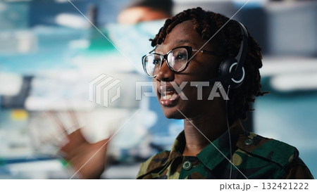 Close up of army telecommunications operator uses tablet and headset to manage secure military communications. Soldier coordinates mission support from command center with surveillance technology Close up of army telecommunications operator uses tablet and headset to manage secure military communications. Soldier coordinates mission support from command center with surveillance technology 132421222