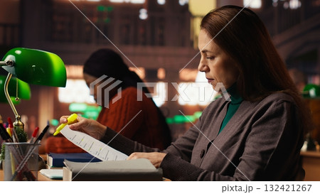 Female person with a focused expression highlighting important ideas from class notes, reading files at college library. Academic research to complete her degree. Self growth. Female person with a focused expression highlighting important ideas from class notes, reading files at college library. Academic research to complete her degree. Self growth. 132421267