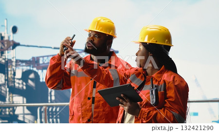 Offshore platform employees supervising deck operations with device, speaking on walkie talkie. African american engineers doing maintenance on drilling rig gear, transmitting radio message Offshore platform employees supervising deck operations with device, speaking on walkie talkie. African american engineers doing maintenance on drilling rig gear, transmitting radio message 132421290