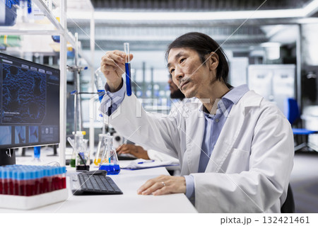 Clinical laboratory technologist investigates chemical sample properties using test tube in laboratory. Asian research facility professional holds vial containing new compound, doing pharmaceutical RD Clinical laboratory technologist investigates chemical sample properties using test tube in laboratory. Asian research facility professional holds vial containing new compound, doing pharmaceutical RD 132421461
