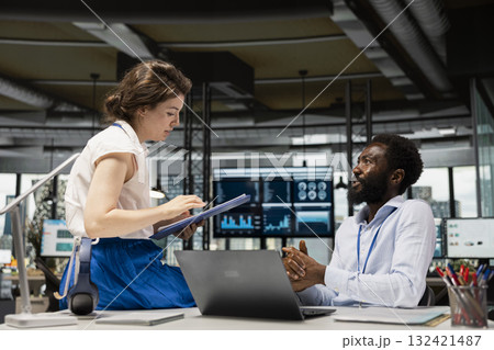 Business partners in multinational office using paperwork and laptop to analyze data. Female executive with clipboard speaking to employee, presenting task updates on budget planning. Business partners in multinational office using paperwork and laptop to analyze data. Female executive with clipboard speaking to employee, presenting task updates on budget planning. 132421487