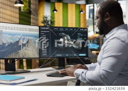 Technician in office analyzes wind turbine component to improve alternative energy efficiency. African american man uses CAD software to refine turbine part design, boosting clean energy output 132421639
