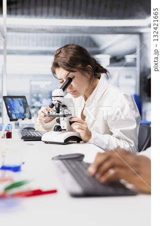 Cytotechnologist evaluates cellular structures using microscope in lab workspace. Technician observes specimens through magnification tool at laboratory bench workstation 132421665