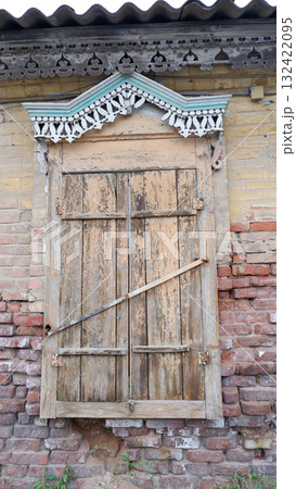 Closed old wooden shutters with rustic carved designs, with intricate carvings at the top. Shutters are antique and installed against a brick wall background. Closed old wooden shutters with rustic carved designs, with intricate carvings at the top. Shutters are antique and installed against a brick wall background. 132422095