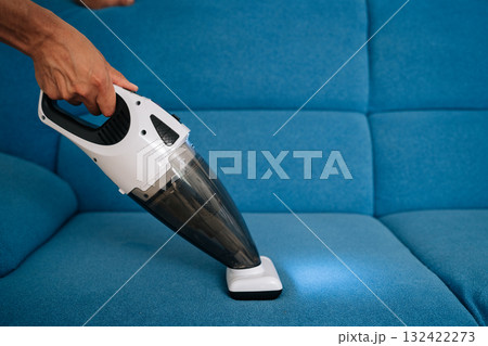 Close-up cropped shot of cleaning service employee using handheld vacuum cleaner to remove dust and dirt from blue sofa, emphasizing importance of cleanliness and hygiene in home maintenance. 132422273