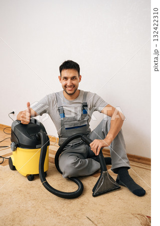Friendly cleaning service employee in overall showing thumbs up gesture sitting on floor with washing vacuum cleaner, ready for thorough floor cleaning and sanitizing service, looking at camera. Friendly cleaning service employee in overall showing thumbs up gesture sitting on floor with washing vacuum cleaner, ready for thorough floor cleaning and sanitizing service, looking at camera. 132422310