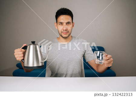 Portrait of young man sitting at table, holding kettle and glass cup, preparing to brew drip coffee in modern kitchen room with minimalist decor, looking camera. Concept of enjoying morning ritual. Portrait of young man sitting at table, holding kettle and glass cup, preparing to brew drip coffee in modern kitchen room with minimalist decor, looking camera. Concept of enjoying morning ritual. 132422313