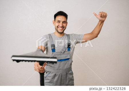 Studio portrait of friendly professional cleaning service employee in overall holding vacuum cleaner and showing thumbs up gesture, smiling looking at camera and recommending cleaning company services 132422326