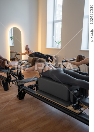 Group of caucasian women exercising with trainer on pilates reformers in a bright studio. Their workout focuses on core strength and flexibility in a modern fitness environment Group of caucasian women exercising with trainer on pilates reformers in a bright studio. Their workout focuses on core strength and flexibility in a modern fitness environment 132422530