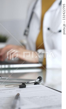 Stethoscope and clipboard with medical documents are on the table near female doctor working on laptop, managing patient data in clinic. Concept of medicine, healthcare and science Stethoscope and clipboard with medical documents are on the table near female doctor working on laptop, managing patient data in clinic. Concept of medicine, healthcare and science 132422705