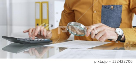 Accountant analyzing financial data with magnifying glass, calculating expenses, and auditing documents at office desk. Audit and taxes in business Accountant analyzing financial data with magnifying glass, calculating expenses, and auditing documents at office desk. Audit and taxes in business 132422706