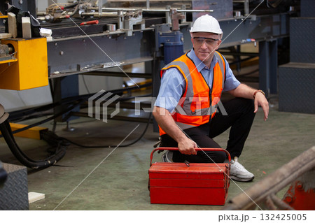 Industrial technician wearing vest safety crouching beside factory machinery with red toolbox, preparing for maintenance. Concept of workplace safety, repair and factory equipment servicing. Industrial technician wearing vest safety crouching beside factory machinery with red toolbox, preparing for maintenance. Concept of workplace safety, repair and factory equipment servicing. 132425005